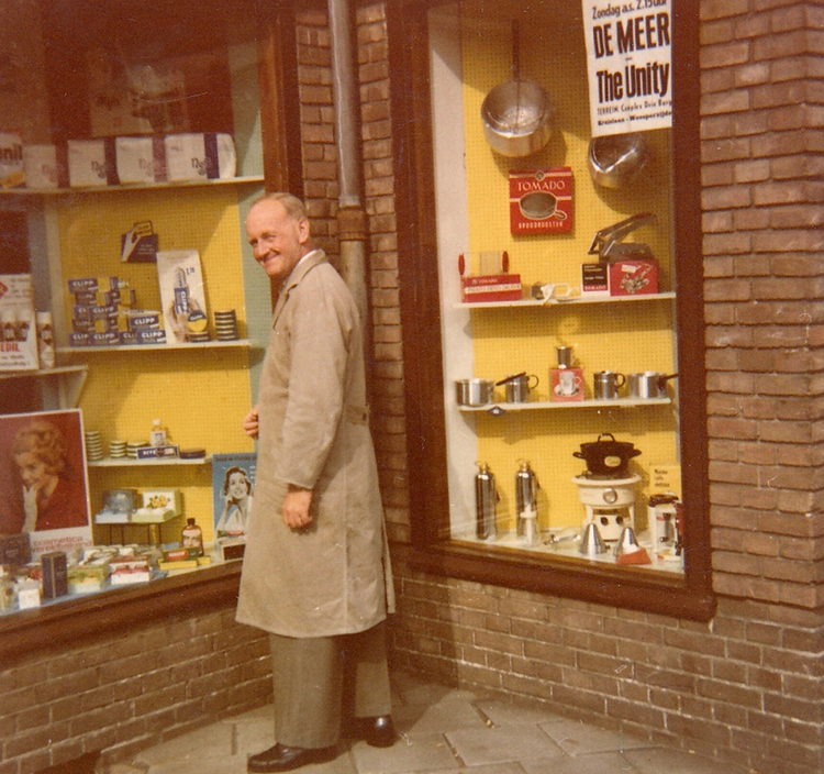 Henk Komen bij de etalage van zijn winkel. - 1963 .<br />Foto: Familie Komen .<br />Aan het raam een aankondiging van de wedstrijd De Meer tegen The Unity -  seizoen 1963/1964<br />Foto: Familie Komen 