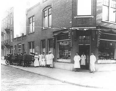 jan van deudekom Bakkerij en winkel van Jan van Deudekom (omstreeks 1930) op de hoek Wakkerstraat / Willem Beukelsstraat. Rechts en links van de winkeldeur staan bakker Jan van Deudekom en zijn vrouw, in de ingang toenmalige winkeljuffrouw Lies van Putten. Op de stoep broodkarren met broodbezorgers Antoon Hoeboer (die hielp 's-nachts ook in de bakkerij), Piet van der Ende en Lucas Lucassen. Daarnaast het bakkerijpersoneel (aan de kleding kan je de functie herkennen). v.l.n.r.: Chef te Deus (lange sloof) deegmaker en algemene leiding; Jan de Roemer (korte sloof) bankman; Anton de Heij (lange sloof en gekleurd truitje) ovenist; banketbakker Klepper (sloof en bakkersbuis); leerling banketbakker in vol ornaat. (Foto en tekst beschikbaar gesteld door J.A.M. van Deudekom) 