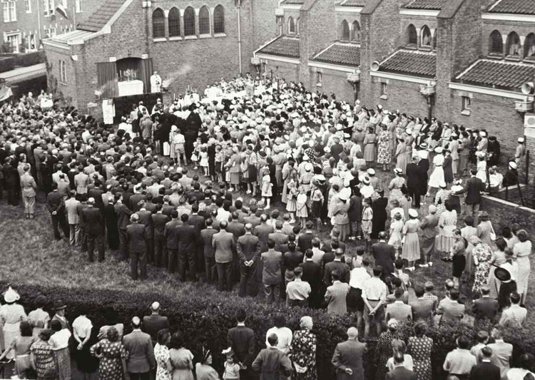 De kerk werd op 21 maart 1929 ingewijd. Op de foto een processie in de jaren 20 op het terrein van de kerk. Eigenlijk was een processie op straat verboden, maar omdat het op eigen terrein gebeurde, kon men dit ongestraft doen. .<br />Foto: Stadsarchief Amsterdam .<br />Foto: Theo Proeskie 