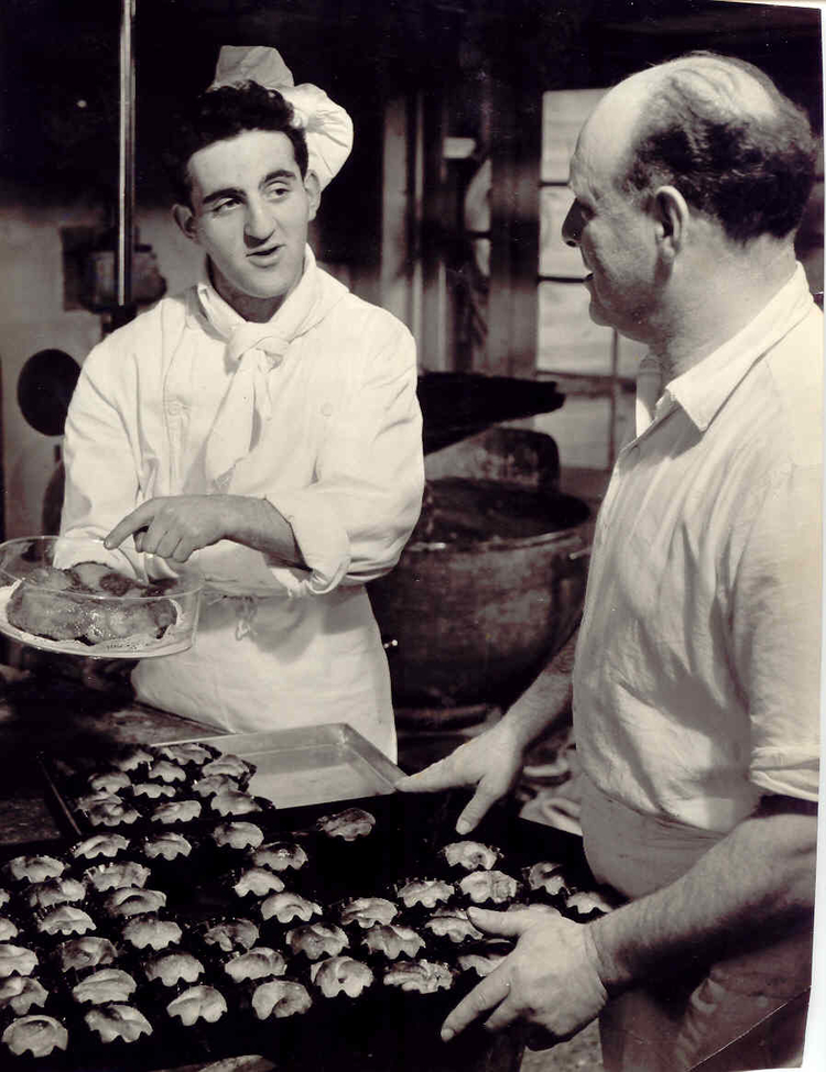 Meijer en David Verdooner in de bakkerij. David Verdooner samen aan het werk in de bakkerij met zijn vader Meijer. De foto is waarschijnlijk gemaakt aan het einde van de jaren vijftig. De foto is afkomstig uit het foto archief van David en Gerda Verdooner 