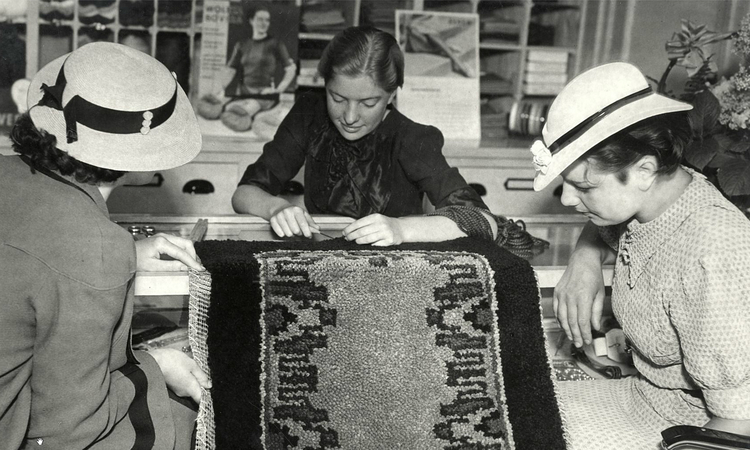 Middenweg 81 - 1937 Twee dames met hoedjes op laten zich door een verkoopster adviseren in het knopen van een wollen tapijt in de wolwinkel van de firma Wernekinck aan de Middenweg te Amsterdam.<br />.<br />Foto: www.gahetna.nl 