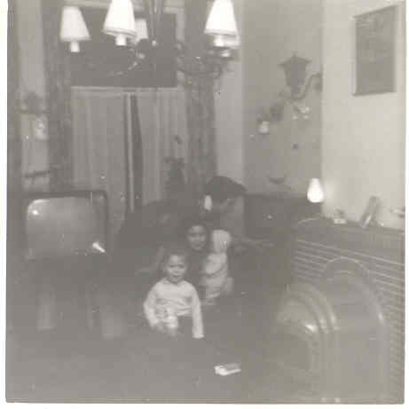 Huiskamer met smeedijzer De huiskamer in 1963 met moeder, broer John en Remine met pop. 'Wij waren een van de eerste families in de straat met televisie. Let op de lampen, mijn vader maakte van alles van smeedijzer.' 