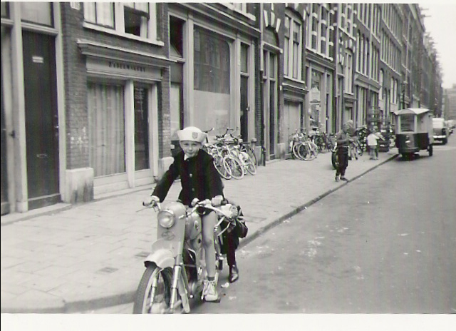 Henny op 10/11-jarige leeftijd (1964 of 1965) in de Derde Oosterparkstraat op de brommer van zijn neef. Zijn broer (bijna onzichtbaar) houdt de brommer vast zodat hij niet omvalt. Op de achtergrond zie je de driewieler van de melkboer Fam. Houweling waarmee hij de buurt door vente.  
