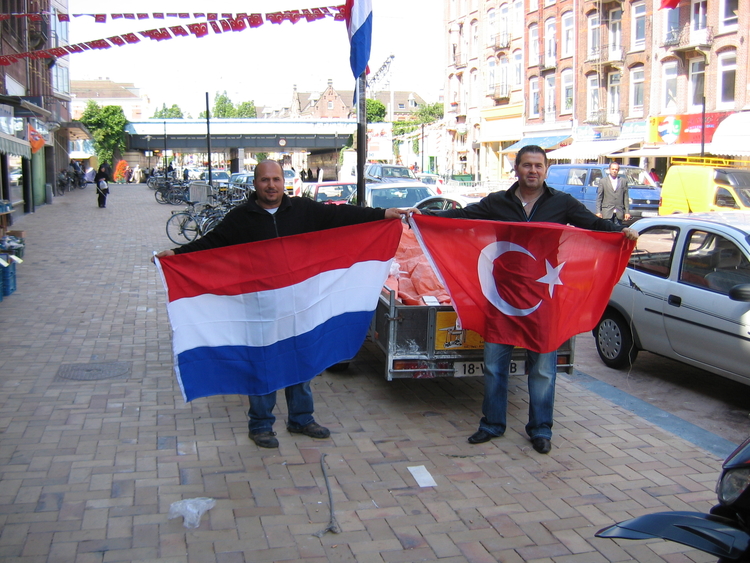 Celal Yildiz (rechts) samen met een medewerker druk bezig met het ophangen van de vlaggen. De foto is gemaakt voor de Yildiz market in de Javastraat, op dinsdag 17 juni. Afgebeeld zijn Celal Yildiz en een medewerker. Zij zijn druk met het ophangen van de Turkse en de Nederlandse vlaggen. De foto is gemaakt door de schrijver. 