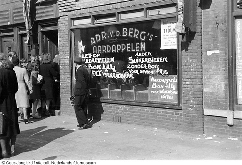 Er is nieuwe voorraad! Voor een groentewinkel met nieuwe voorraden aardappelen en suikerbieten staat een rij klanten, Amsterdam (mei 1945). Bron: Geheugen van Nederland, fotograaf Cees Jongkind.  