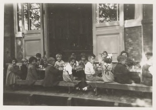 Kinderen eten buiten tijdens de middagpauze op de speelplaats achter de Sophie Rosenthal Bewaarschool. Adres: Nieuwe Uilenburgerstraat 27-31, september 1940. Bron: fotocollectie SAA.  